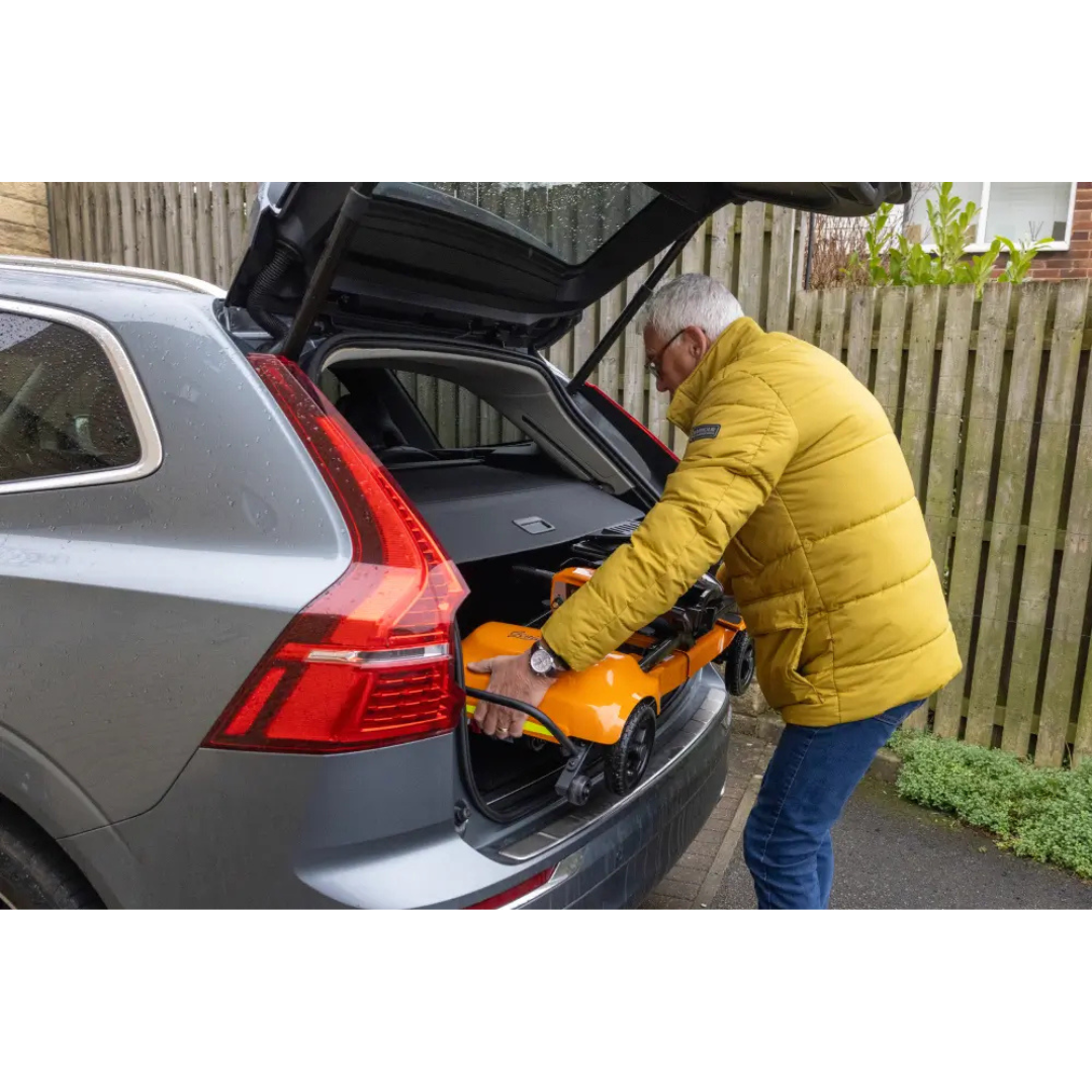 A man in a yellow jacket loads a folded Monarch Genie Ultralight Portable Mobility Scooter into the trunk of a silver SUV parked near a wooden fence.