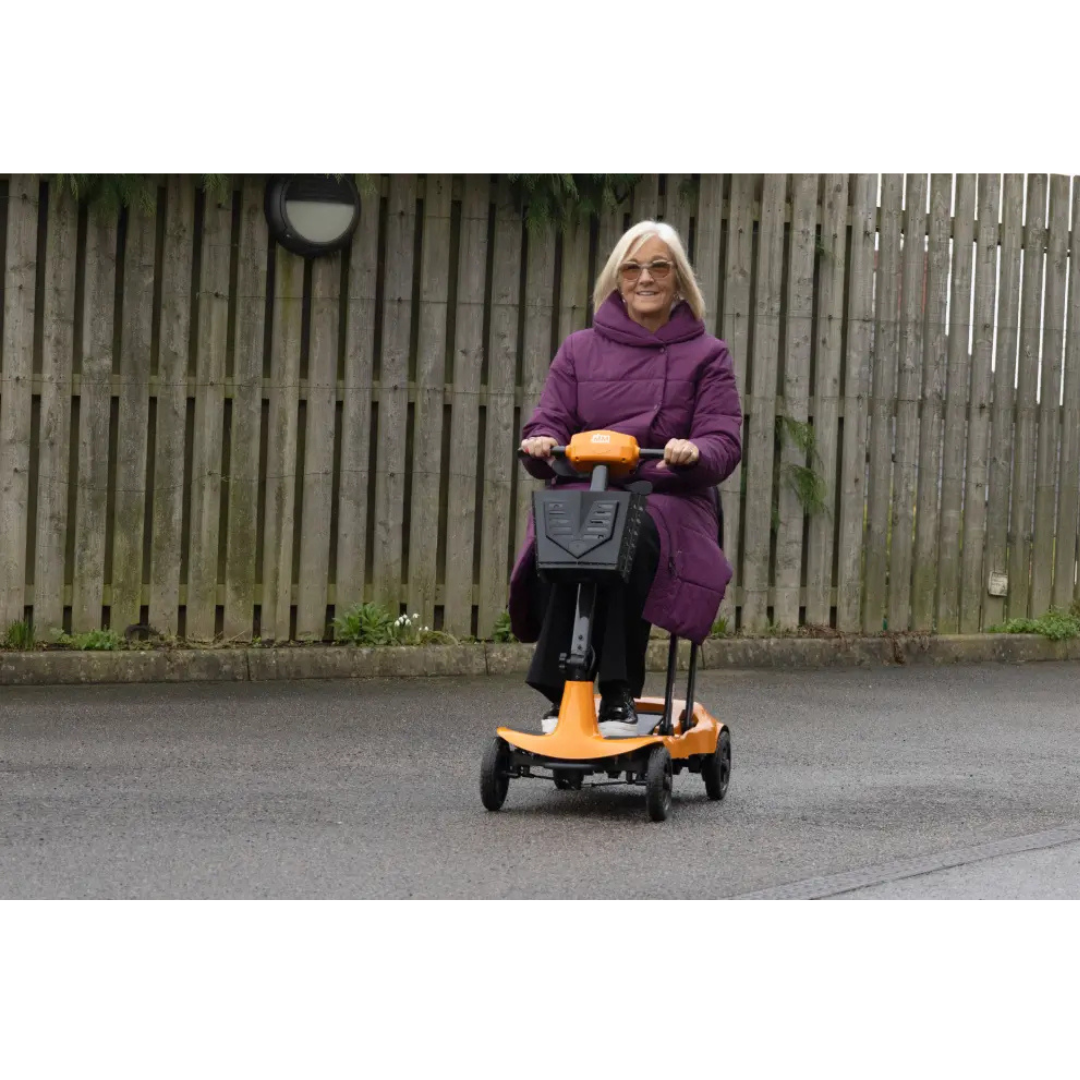 An older woman in a purple coat rides the Monarch Genie Ultralight Portable Mobility Scooter on a paved path outdoors, with a wooden fence and greenery behind her—perfect for showcasing this mobility aid in marketing materials.