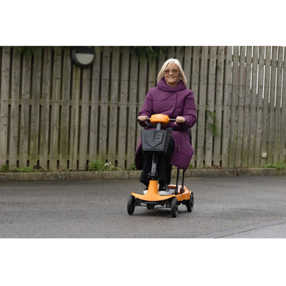 An older woman in a purple coat rides the Monarch Genie Ultralight Portable Mobility Scooter on a paved path outdoors, with a wooden fence and greenery behind her—perfect for showcasing this mobility aid in marketing materials.