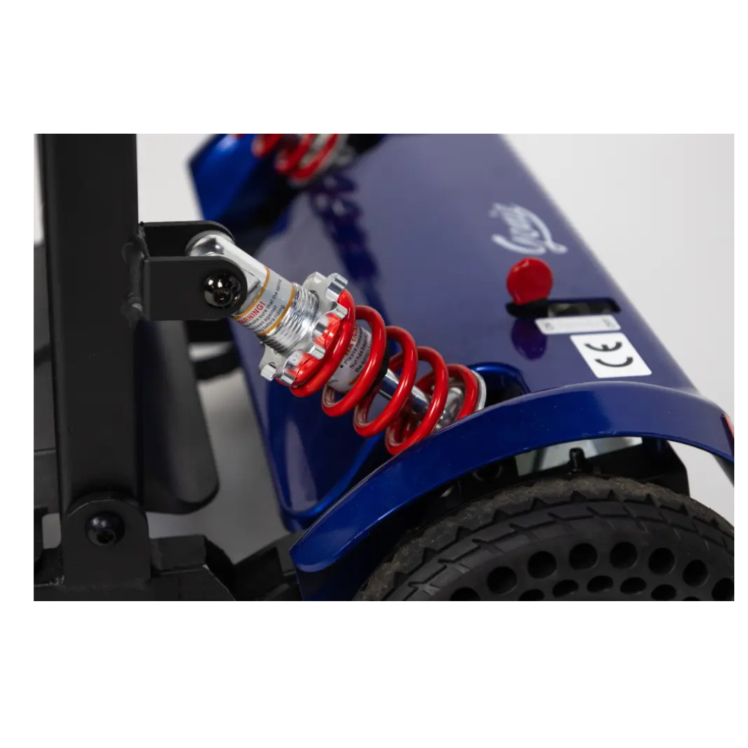 Close-up of the Monarch Genie Lightweight Portable Scooter’s blue rear wheel, featuring a red suspension spring, shock absorber, black treaded tire with holes, and a CE certification label—ideal for Monarch Genie SEO keywords.