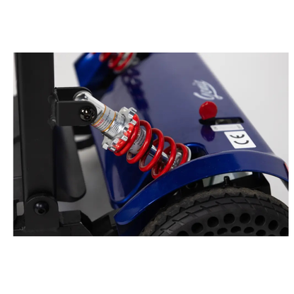 Close-up of the Monarch Genie Lightweight Portable Scooter’s blue rear wheel, featuring a red suspension spring, shock absorber, black treaded tire with holes, and a CE certification label—ideal for Monarch Genie SEO keywords.