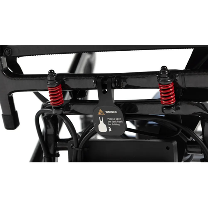 Close-up of a bicycle frame showing two red suspension springs and a warning label instructing to open two lock hooks for folding—similar to the practical mechanisms found on ultra-lightweight folding mobility devices such as the Monarch MM60 Carbon Ultralite Electric Wheelchair. Black wires are visible near the locking mechanism.