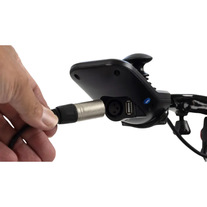 A hand is plugging a connector into a black electronic device mounted on the handlebar of the Monarch MM60 Carbon Ultralite Electric Wheelchair, which features a USB port and a circular input port.