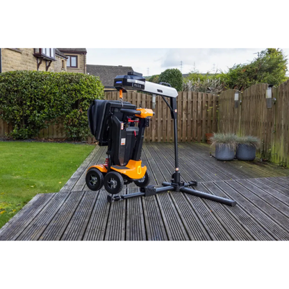 A black and orange mobility scooter is parked on a wooden deck in a lush, fenced garden next to the Monarch Atlas 4 Portable Car Lift - Mobility Aid Lifter, which features a sturdy metal arm.