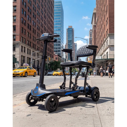 A Monarch Supa Carbon Lite Ultra-Lightweight Carbon-Fiber Scooter with four wheels and two seats is parked on a city sidewalk, bordered by tall buildings, yellow taxis, and pedestrians under the sunny sky.