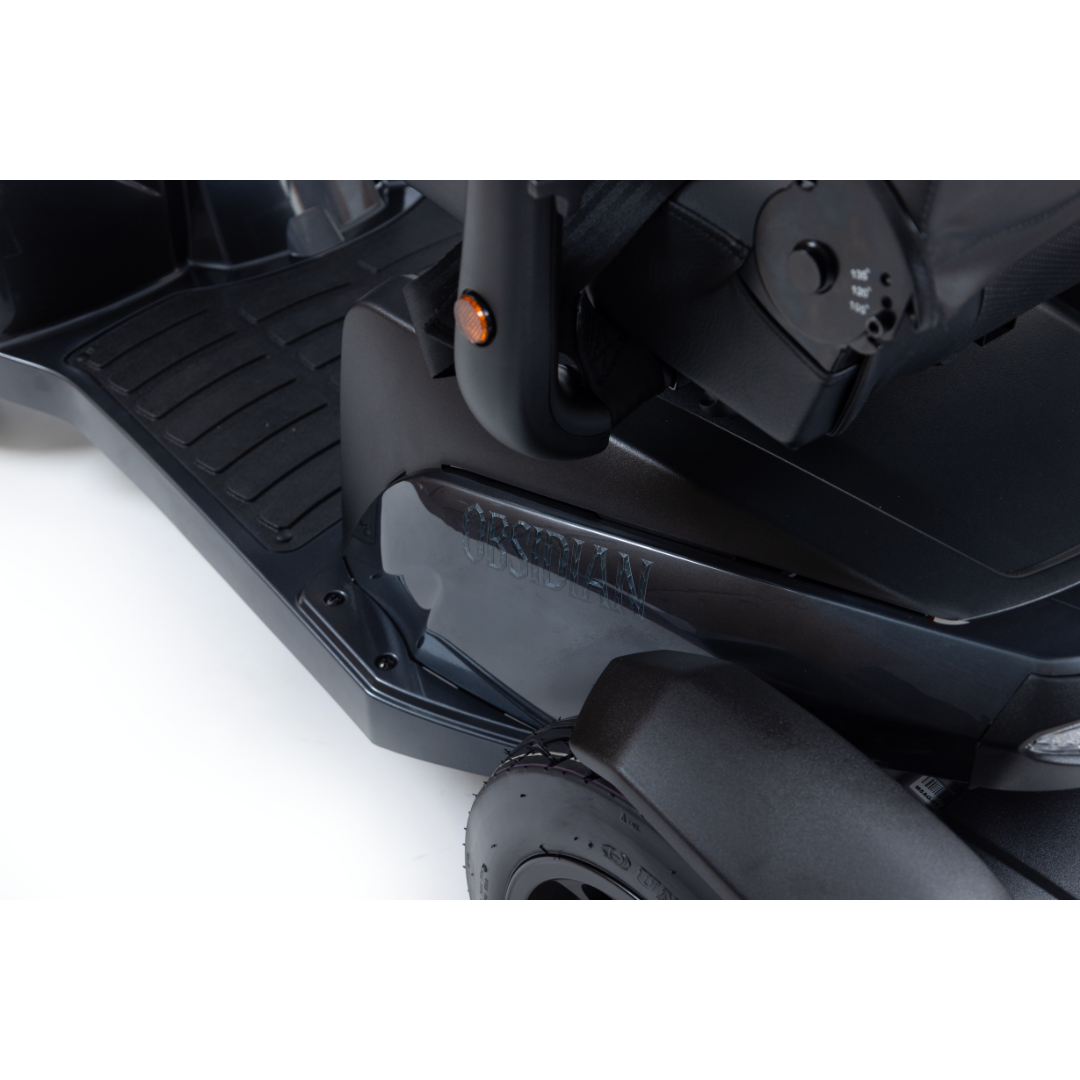 Close-up of the base and footrest area of a dark-colored Monarch Obsidian HD Outdoor Mobility Scooter, showing part of the seat, wheel, and side panel label. Check specifications or watch videos for more details.