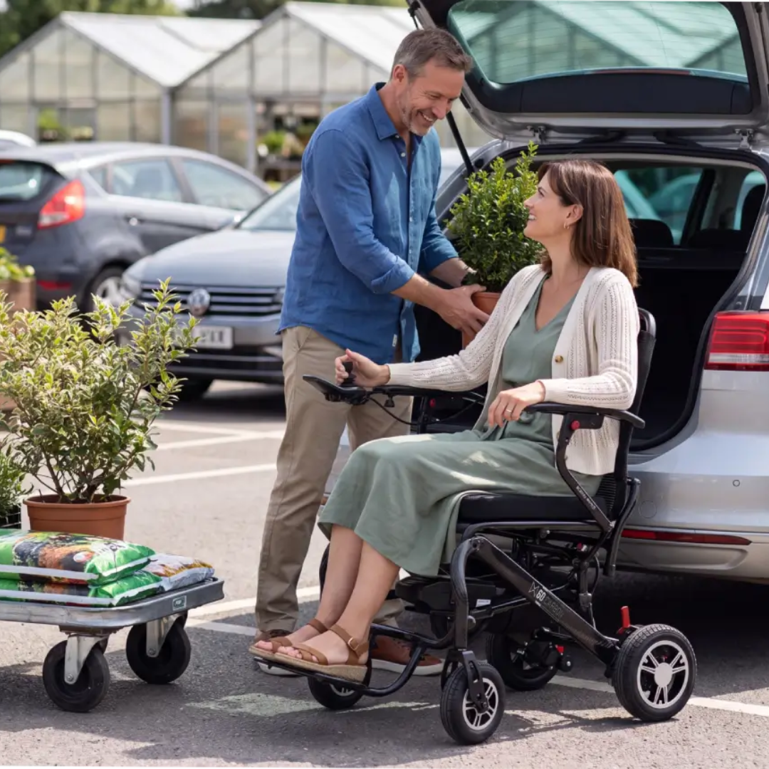 A woman in a Monarch MM60 Carbon Ultralite Electric Wheelchair sits by an open car trunk, smiling at a man who is unloading a potted plant. Gardening supplies and plants are on a cart nearby in a parking lot.