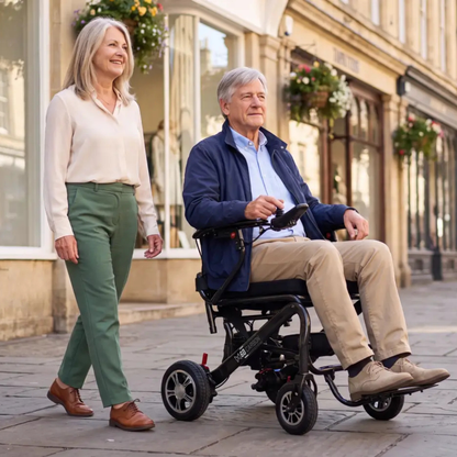 An older man in a Monarch MM60 Carbon Ultralite Electric Wheelchair and an older woman walk together on a sunny city sidewalk, smiling, with shops and hanging flowers visible in the background.