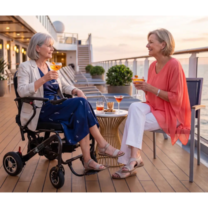 Two older women relax on a cruise ship deck, one comfortably seated in a Monarch MM60 Carbon Ultralite Electric Wheelchair, as they enjoy cocktails and smile at each other near a small table, with the sun setting over the ocean.