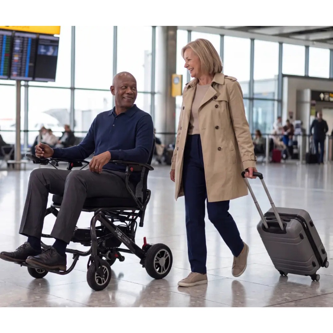 A man in a Monarch MM60 Carbon Ultralite Electric Wheelchair and a woman with a suitcase smile at each other while moving through an airport terminal.