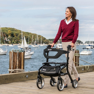 A smiling woman in a red sweater and beige pants uses the Drive Nitro® Sprint Rollator with Accessory Bundle on a wooden dock, with sailboats and trees in the background.