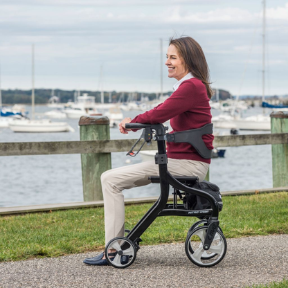 A woman in a red sweater and beige pants rides a black mobility scooter along a waterfront path, smiling and relaxed, with her Drive Nitro® Sprint Rollator with Accessory Bundle by her side as boats and water appear in the background.