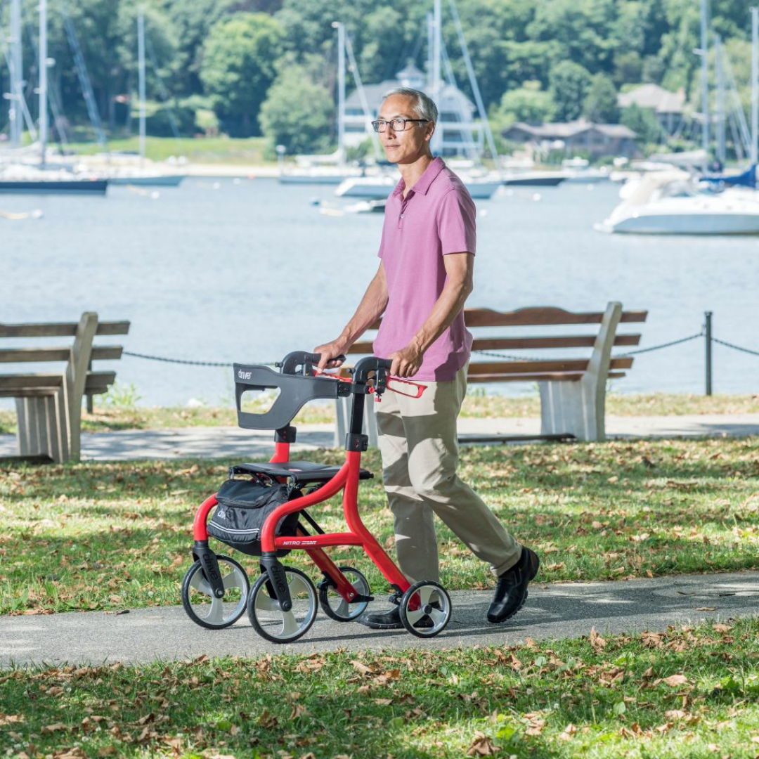 An older man in glasses, a pink polo, and khaki pants walks with the Drive Nitro® Sprint Rollator with Accessory Bundle along a park path by the water, with boats and trees visible in the background.