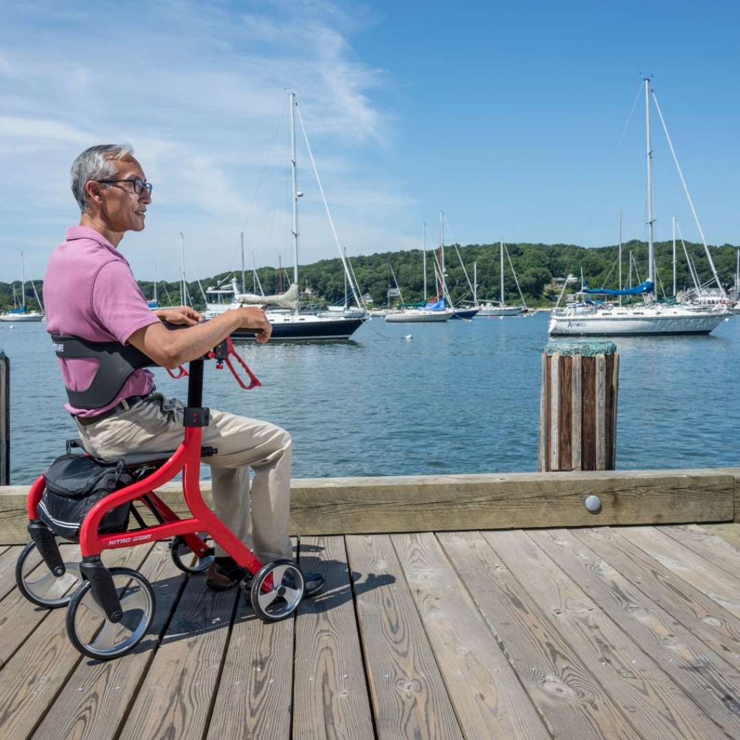 An older man sits on a wooden dock by the water with his Drive Nitro® Sprint Rollator with Accessory Bundle, watching sailboats anchored in the sunny harbor as green trees and blue sky create a peaceful backdrop.