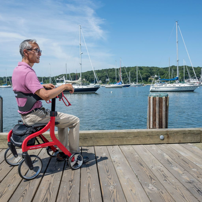 An older man sits on a wooden dock by the water with his Drive Nitro® Sprint Rollator with Accessory Bundle, watching sailboats anchored in the sunny harbor as green trees and blue sky create a peaceful backdrop.