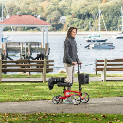 A woman with a leg injury smiles while using the Drive Folding Nitro® Glide Knee Walker—an alternative to crutches—in a waterfront park with boats and trees. She wears a gray jacket and light pants.