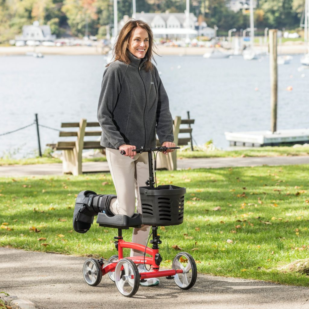 A woman with a leg in a medical boot smiles while using the Drive Folding Nitro® Glide Knee Walker, a popular crutches alternative, on a paved path near a waterfront with benches, grass, and boats in the background.