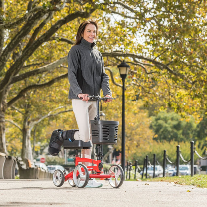 A woman with a medical boot on her right leg smiles as she rides a red Drive Folding Nitro® Glide Knee Walker, a popular crutches alternative, along a tree-lined park path with benches on a sunny day.