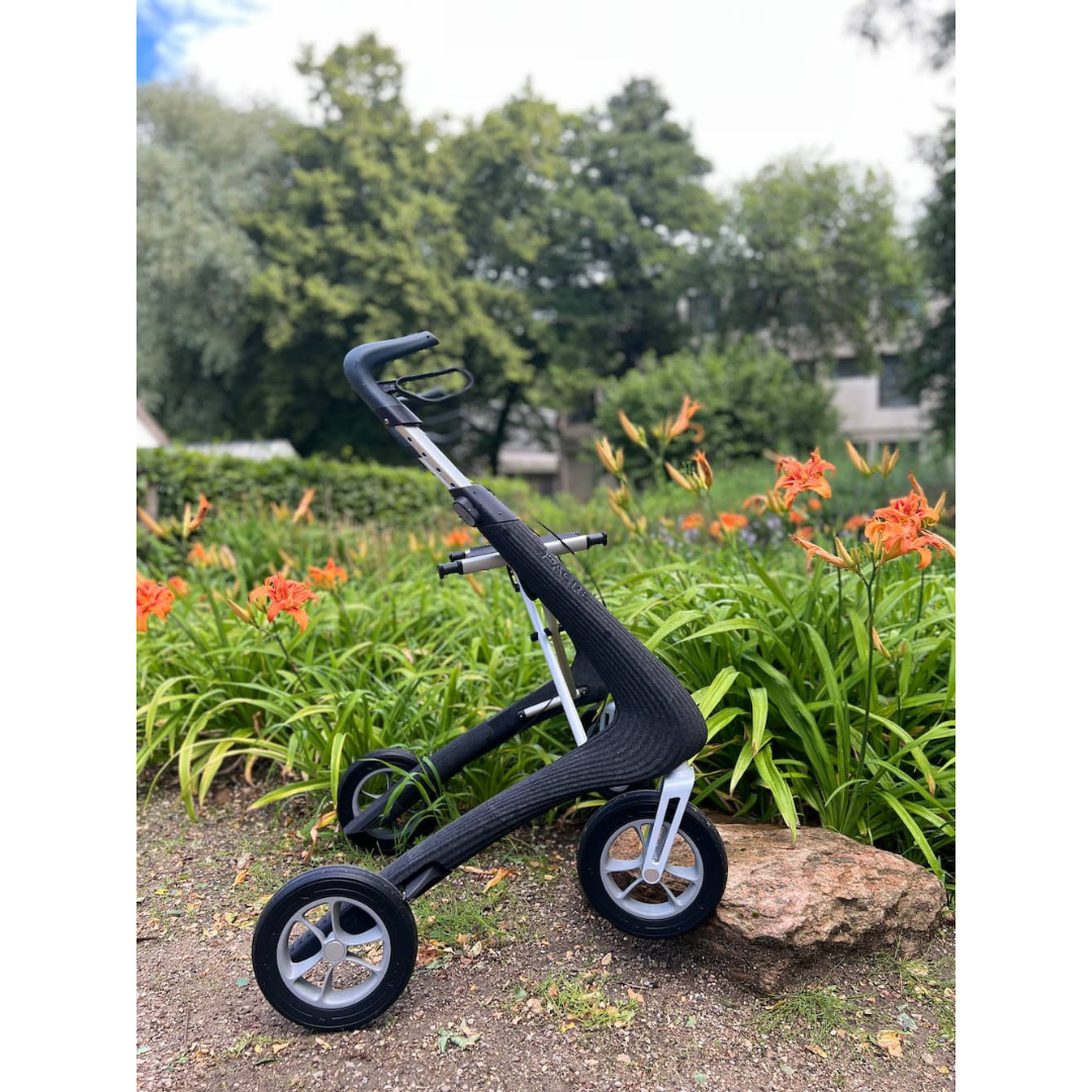 A byACRE Nordic Pioneer Lightweight Folding Rollator - Only 13 lbs in sleek black with three wheels is parked on a garden path near orange lilies and green foliage, with trees and a cloudy sky in the background.