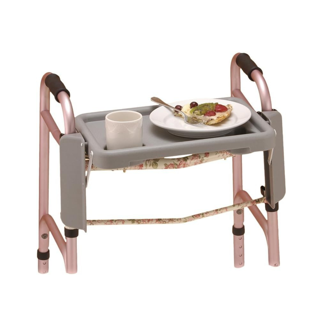 A pink NO Walker features the Nova Folding Walker Tray with Two Cup Holders attached, holding a white mug and a plate of food. It also has black hand grips and floral-patterned padding on the crossbar.