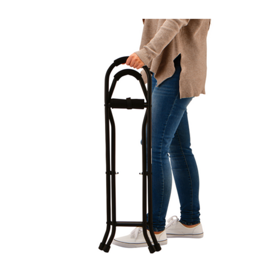 A person holds the handle of a folded black Nova 3092 Folding Travel Seat Cane With Sling Seat, wearing a beige sweater, blue jeans, and white shoes. The image is set against a white background.