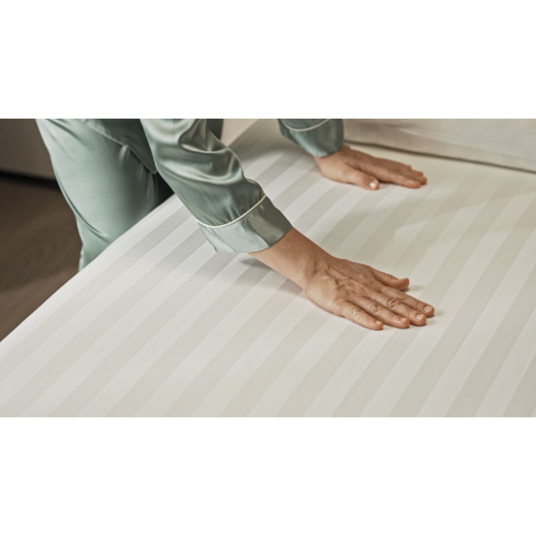 A person in a green satin pajama top is smoothing the surface of a Starsleep Rotating Bed, adorned with crisp, white striped Starsleep Orin Bed Sheets and Protectors. Their hands press down gently to create a tidy and wrinkle-free appearance.