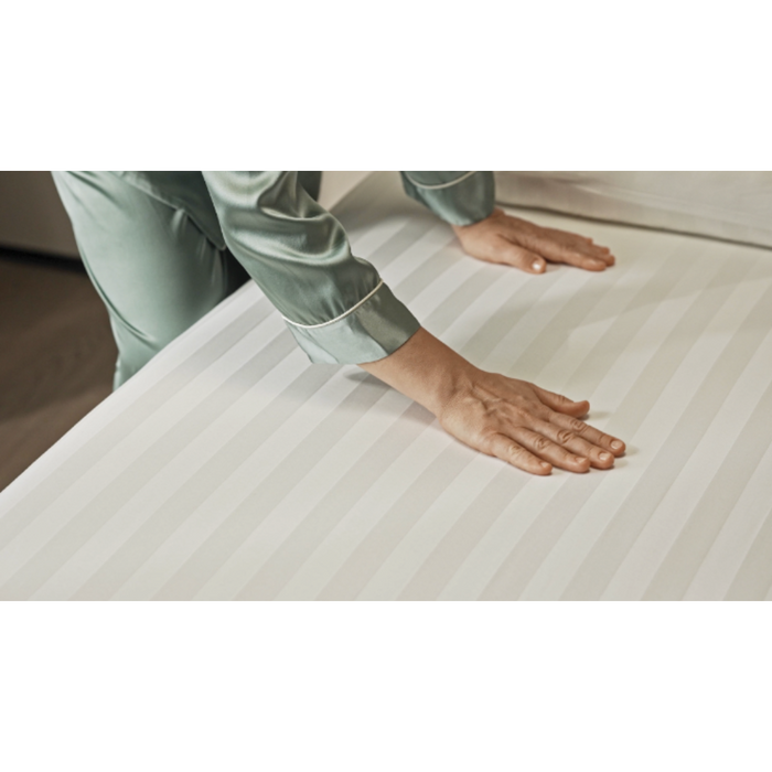 A person in a green satin pajama top is smoothing the surface of a Starsleep Rotating Bed, adorned with crisp, white striped Starsleep Orin Bed Sheets and Protectors. Their hands press down gently to create a tidy and wrinkle-free appearance.