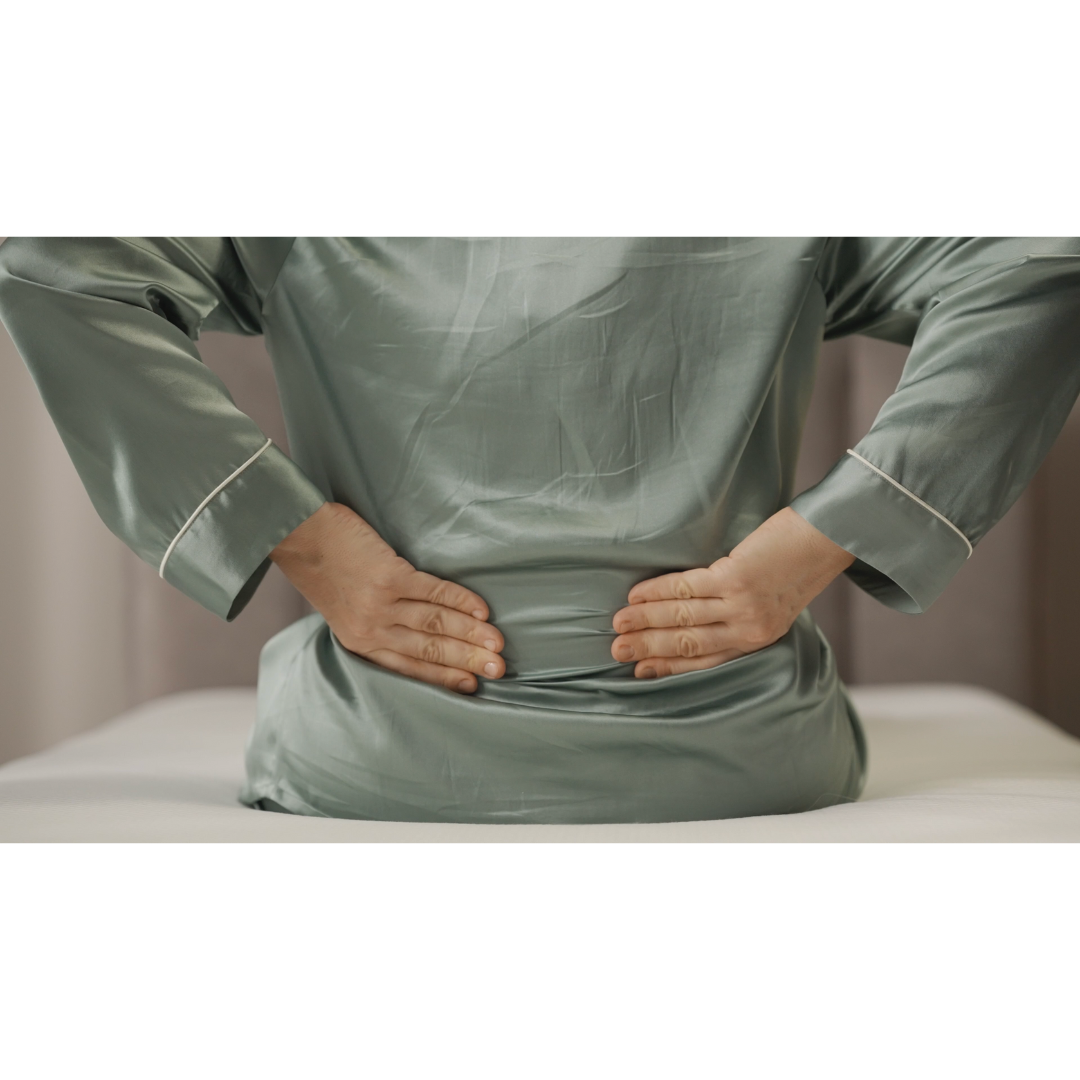 A person in green silk pajamas sits on a Starsleep Orin Rotating Full Electric Lift Assist Bed with Heat & Massage, pressing their hands against their lower back. The warmly blurred room hints at serenity as the multifunctional bed offers support.