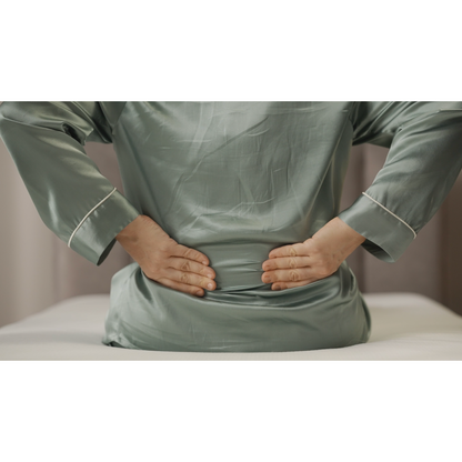 A person in green silk pajamas sits on a Starsleep Orin Rotating Full Electric Lift Assist Bed with Heat & Massage, pressing their hands against their lower back. The warmly blurred room hints at serenity as the multifunctional bed offers support.