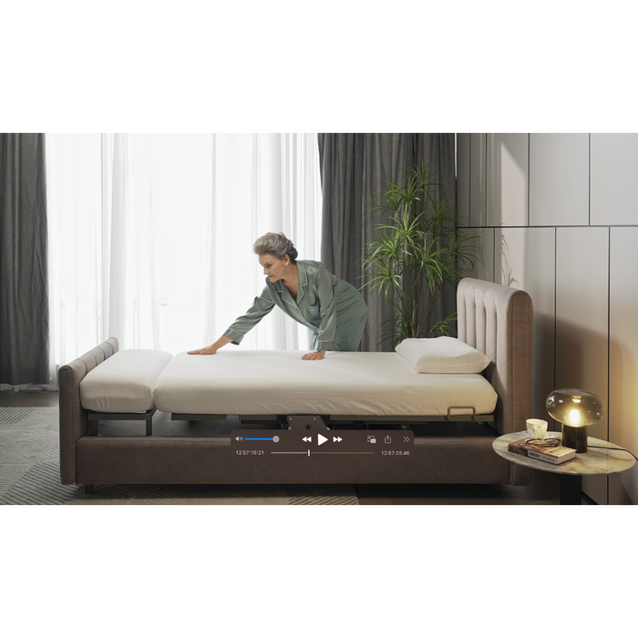 Dressed in green pajamas, someone makes a bed in a modern room featuring the plush Starsleep Orin Rotating Full Electric Lift Assist Bed with heat and massage. A round table with a lamp and plant adds decor, while sunlight through open curtains highlights an on-screen video player bar.