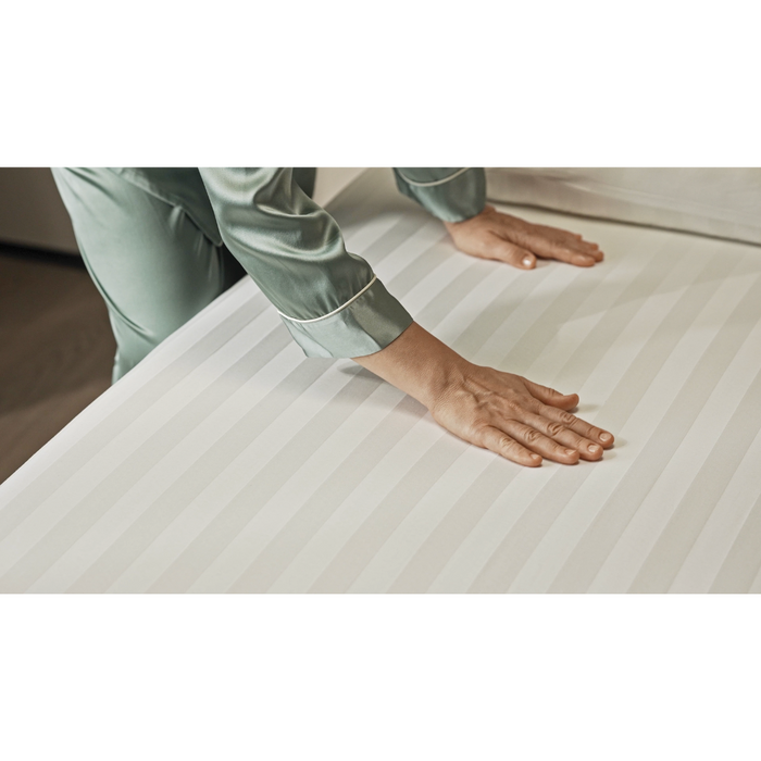 Wearing green pajamas, a person smoothes a white striped bedsheet on the Starsleep Orin Rotating Full Electric Lift Assist Bed with Heat & Massage. The softly lit room highlights the fabrics subtle texture and sheen.