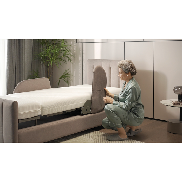 An older woman in a light green outfit and slippers kneels by the Starsleep Orin Rotating Full Electric Lift Assist Bed with Heat & Massage to examine its silent German motor. The modern room includes a potted plant, round side table, and window curtain.