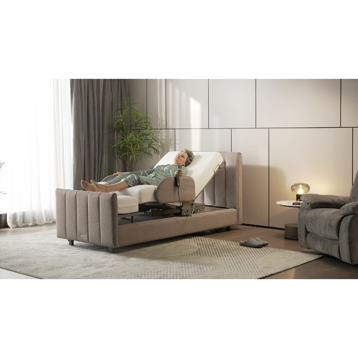 In a modern bedroom, a person in pajamas relaxes on the Starsleep Orin Rotating Full Electric Lift Assist Bed with Heat & Massage, its head elevated silently. A nearby small table with a lamp, reclining chair, plant, and soft rug enhance the space as light streams through window curtains.