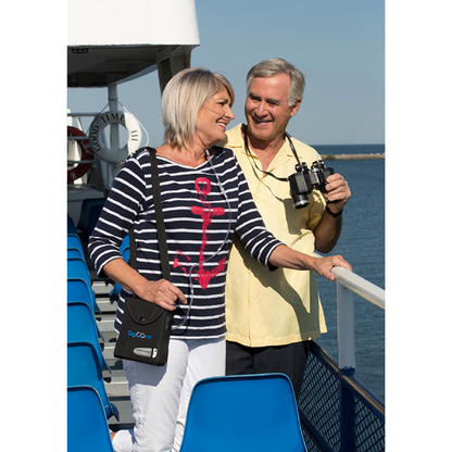 A smiling older couple stands on a boat deck by the water; the woman wears a striped shirt with a red anchor and carries a black bag, while the man holds binoculars and their lightweight OxyGo FIT Compact Portable Oxygen Concentrator.