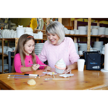 An older woman using an OxyGo FIT Compact Portable Oxygen Concentrator and a young girl smile while decorating a ceramic cupcake at a table with paintbrushes and art supplies in a pottery studio, with shelves of white ceramics in the background.