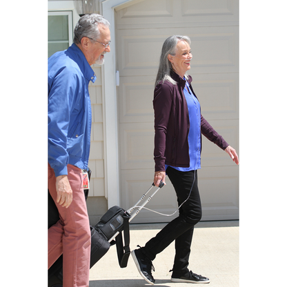 An older woman, casually dressed, smiles while walking outside with a black rolling suitcase and her OxyGo NEXT Ultralite Portable Oxygen Concentrator, beside an older man, as they pass a beige garage door on a sunny day.