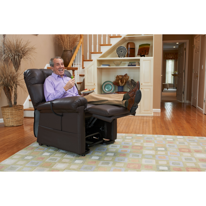 An older man sits comfortably in a Golden Tech PR510 MaxiComfort Cloud Recliner with Lift Assist - Extra Wide, enjoying a snack. He wears a purple shirt and beige pants. The living room features wooden floors, a patterned rug, and a staircase in the background.