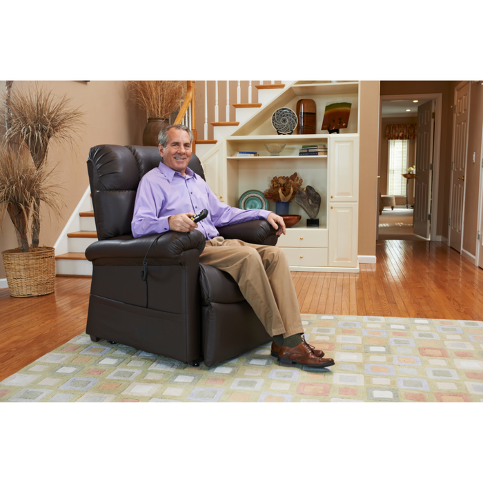 A person relaxes in a Golden Tech PR510 MaxiComfort Cloud Recliner with Lift Assist - Extra Wide, holding a remote control in the cozy living room. Wooden floors and a patterned rug complement the staircase adorned with decorative items in the background.