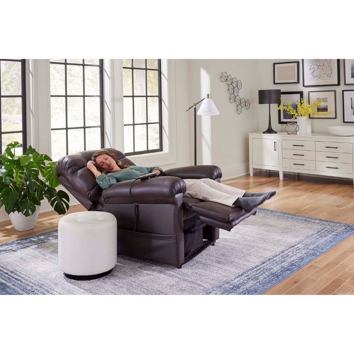 A person is peacefully sleeping in a Golden Tech PR510 MaxiComfort Cloud Recliner with Lift Assist - Extra Wide, featuring Zero Gravity+ positioning, in a bright living room. Sunlight from large windows illuminates the white ottoman, floor lamp, and sideboard with decor. A large plant sits on a blue and white rug next to the chair.