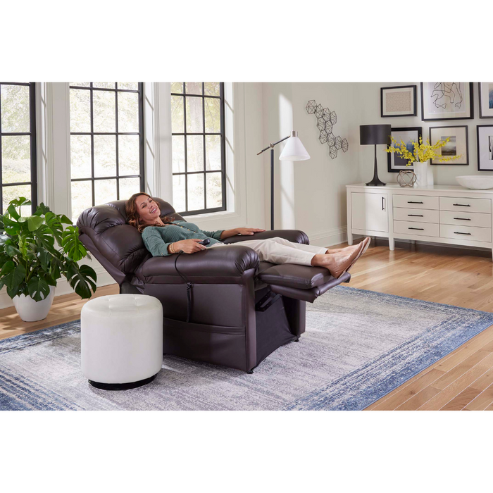 In a bright living room with large windows, a blue area rug, a white ottoman and sideboard, various wall decorations, and a floor lamp, someone relaxes on the Golden Tech PR510 MaxiComfort Cloud Recliner With Lift Assist - Extra Wide using Zero Gravity+ (ZG+) positioning.