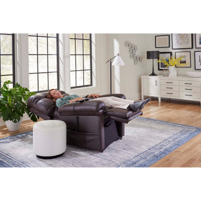 A person relaxes with eyes closed in a bright living room, seated in a Golden Tech PR510 MaxiComfort Cloud Recliner With Lift Assist - Extra Wide by the window. The chairs Zero Gravity+ (ZG+) feature ensures relaxation. A white ottoman, floor lamp, sideboard with decor, and potted plant rest on a blue-bordered rug nearby.