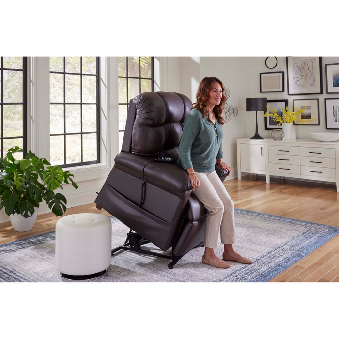 A woman rises from a PR510 MaxiComfort Cloud Recliner with Lift Assist - Extra Wide, in a bright living room. The brown recliner complements large, light-filled windows. A sleek white cabinet is behind her with a plant on the nearby rug.