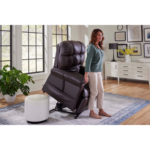 A woman relaxes in a Golden Tech PR510 MaxiComfort Cloud Recliner with Lift Assist - Extra Wide featuring Zero Gravity+ positioning in a bright living room with large windows, a cozy rug, potted plants, framed artwork, and a white ottoman complementing the decor on the sideboard.