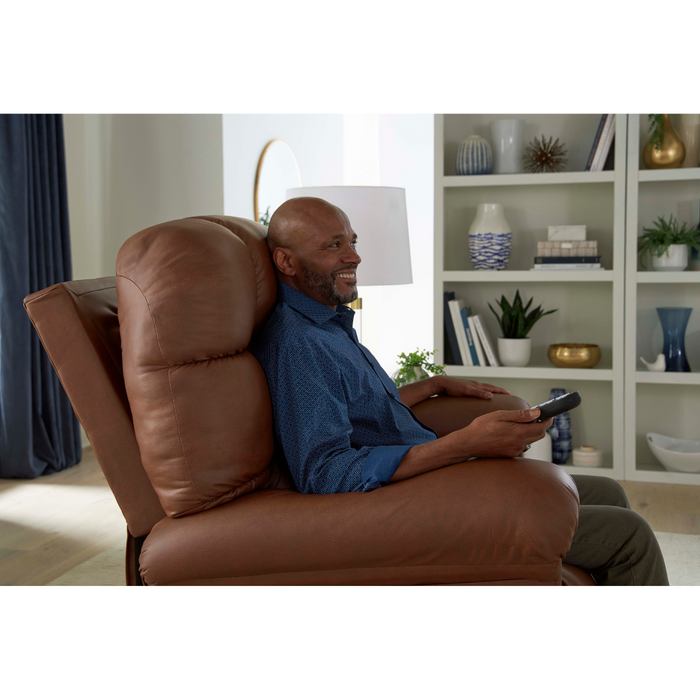 A man relaxes in a Golden Tech PR515 Cloud Recliner With Lift Assist, ZG+ & Twilight - SM/MD, smiling as he holds a remote. A bookshelf with decor, a white lamp, and blue curtains adorn the background while the recliner provides effortless ergonomic comfort.