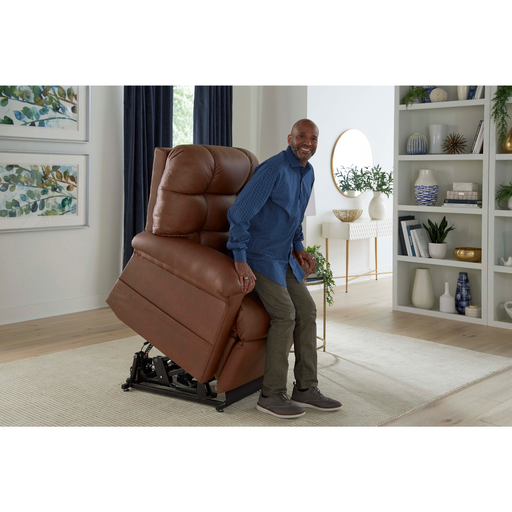 A person rises from a Golden Tech PR515 Cloud Recliner With Lift Assist, ZG+, & Twilight - SM/MD. The room has white walls, a bookshelf with decor, framed wall art, and curtains framing a window near a table with plants.