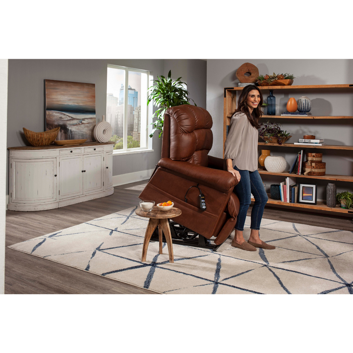 A woman stands next to a brown Golden Tech PR515 Cloud Recliner With Lift Assist in a cozy living room with a white cabinet, wooden bookshelves, coastal artwork, and large windows showing a cityscape. A small wooden table is on a patterned rug beside the chair.