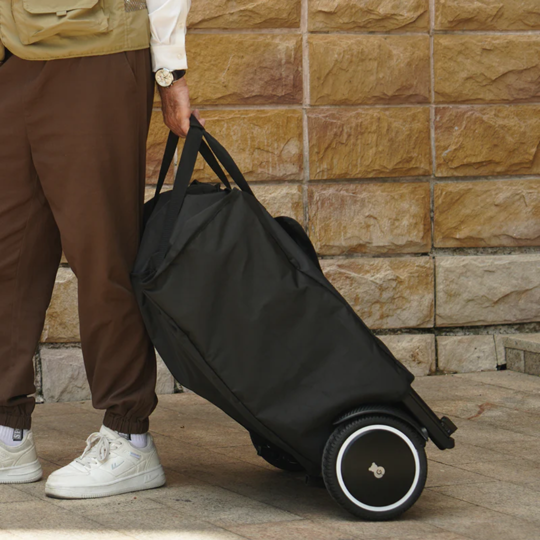 A person wearing brown pants and white sneakers pulls a black Travel Bag For Paiseec Q3 Electric Wheelchair by its handles along a sidewalk in front of a stone wall.