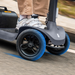 Close-up of a person in white sneakers and light brown pants riding the durable black Paiseec S1 Portable Electric Mobility Scooter, featuring blue-accented wheels for all-terrain performance, smoothly along a street with grassy surroundings in view.