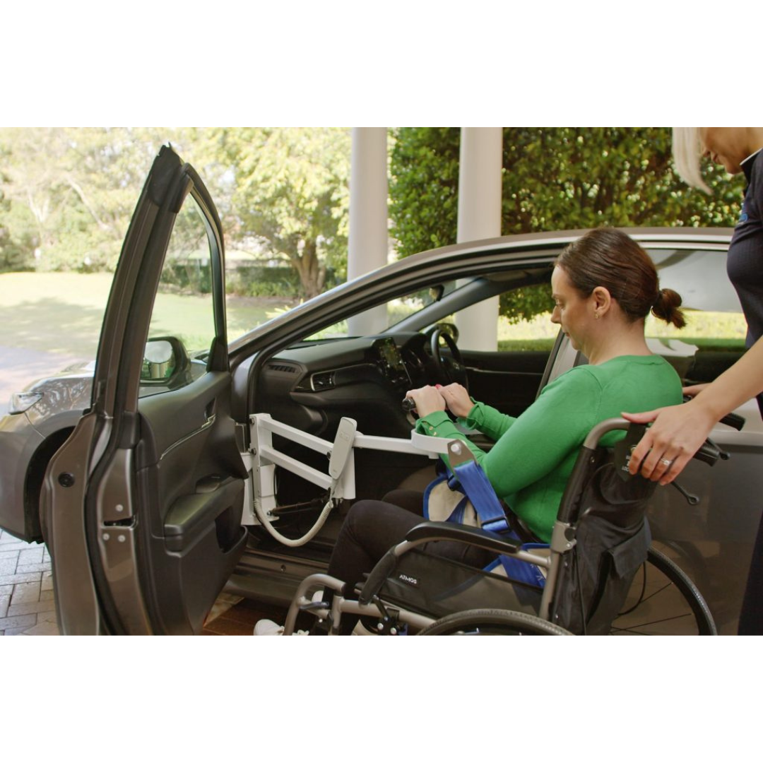 A woman in a green top, seated in a wheelchair, uses the Para Mobility IBIS Wheelchair to Car Access Lift to transfer into the driver’s seat of a car, assisted by another person with this mobility solution.