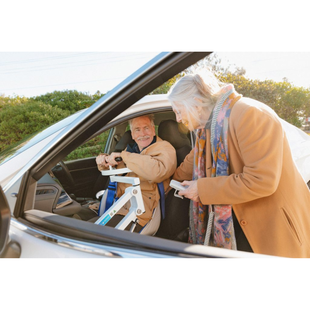 An older man smiles in the driver’s seat as an older woman assists him with a Para Mobility IBIS Wheelchair to Car Access Lift. Both are warmly dressed and appear happy during the vehicle transfer.